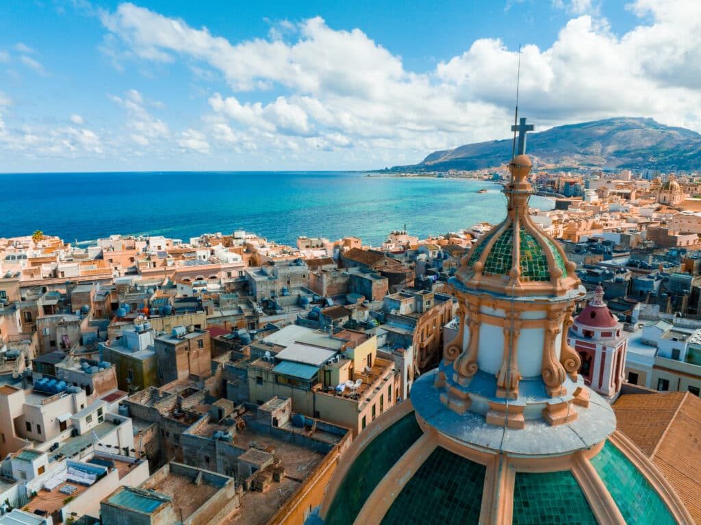 Sicilian coastal city with green-tiled cathedral dome, blue Mediterranean Sea, and rocky mountains.