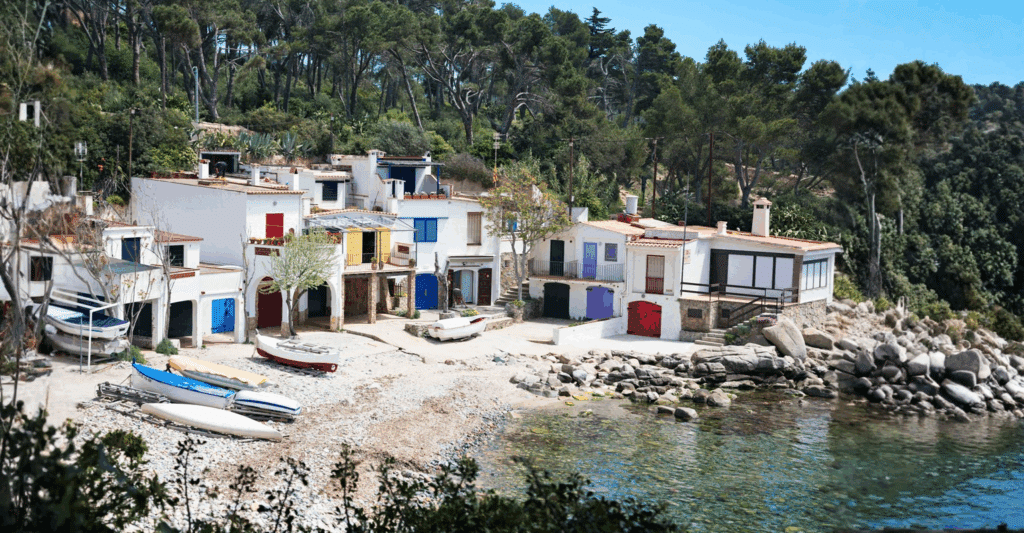 Charming coastal cove with white houses, colorful doors, boats on a pebble beach, backed by pine-covered hills.