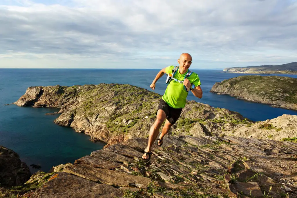 Smiling trail runner in sandals on rugged coastal path with bright blue ocean and distant village.