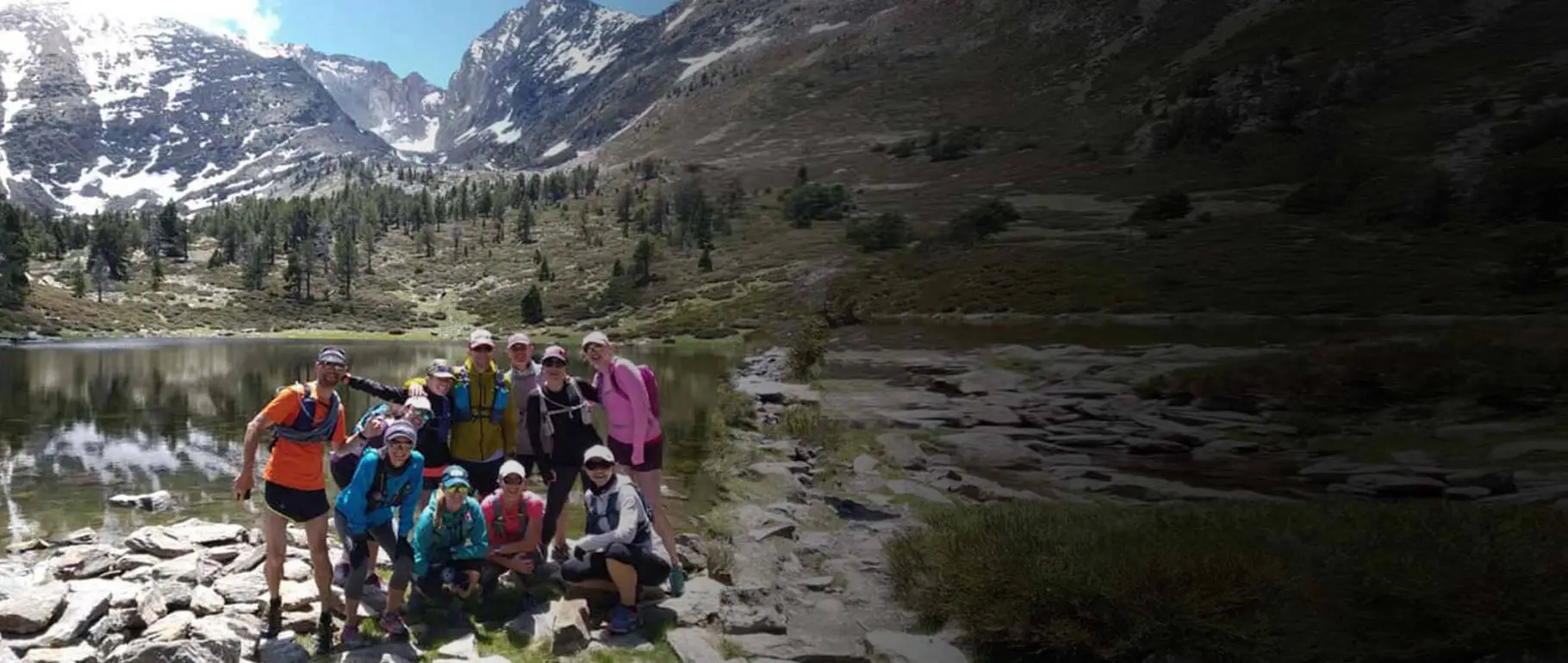 Trail running group poses by a clear mountain lake, with towering snow-capped peaks and pine forests.