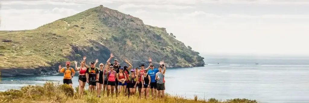 Group of trail runners waving on a coastal path overlooking a rocky mountain and the sea.