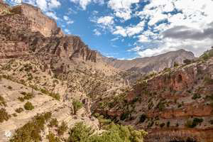 Rugged desert canyon with steep, towering rock formations, sparse green plants, and a winding valley under a blue sky.
