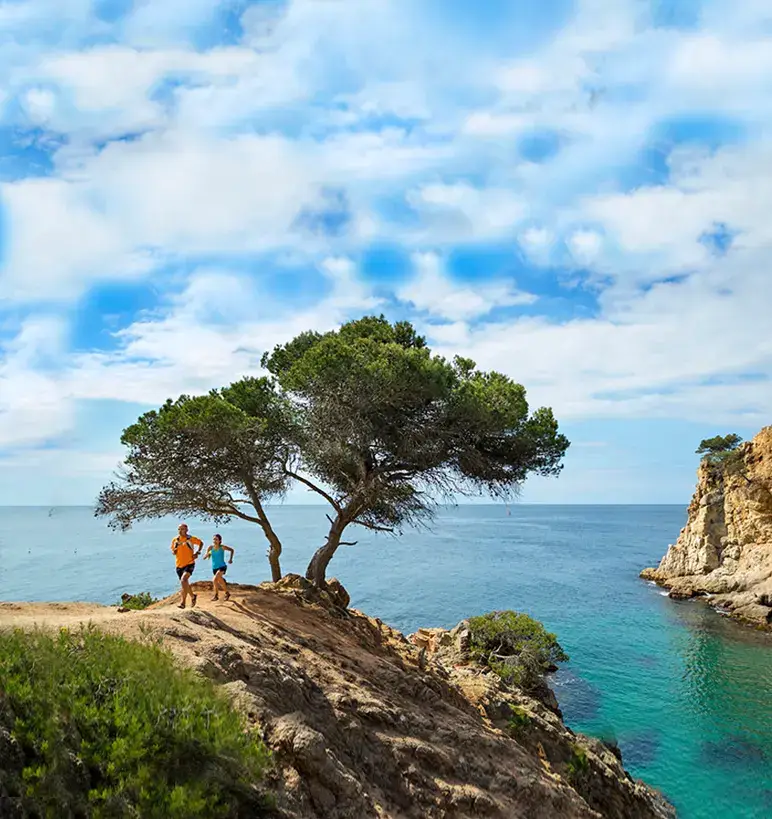Two people trail running on a scenic coastal cliff path overlooking a turquoise ocean and rocky cove.