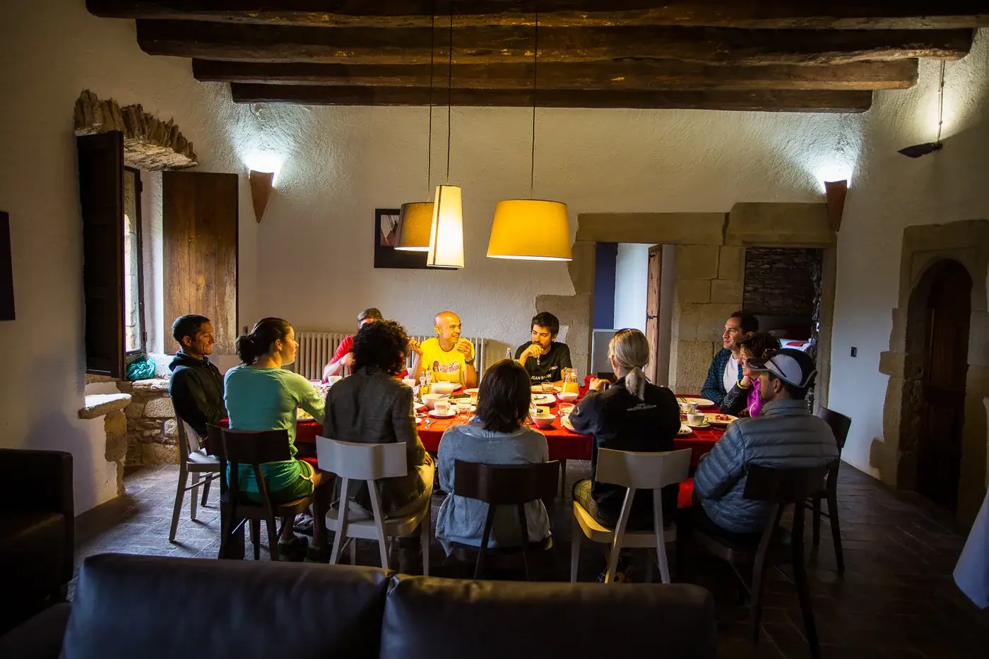 The group sitting around a long table at dinner in a stone Catalan farmhouse, lit by two lamps
