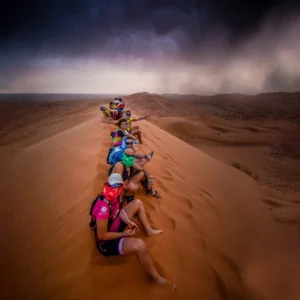 Runners seated in a line down a Sahara dune crest